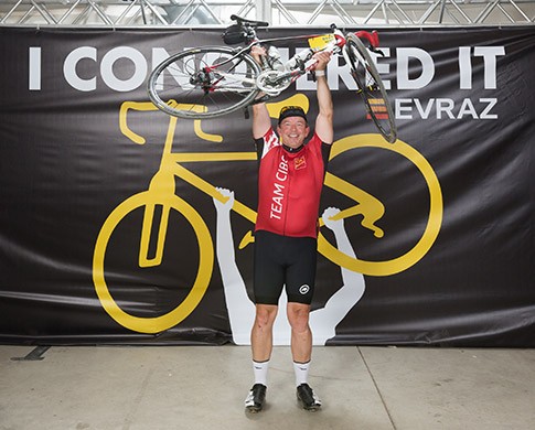 Sean lifts his bike over his head in front of a sign with the Evraz logo reading I conquered it