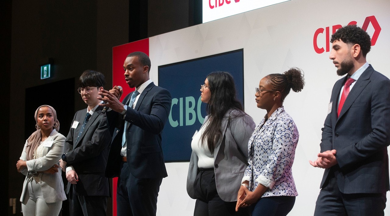 6 students are standing on a stage with the CIBC logo in the background. One student has a microphone and is speaking while the others look on.