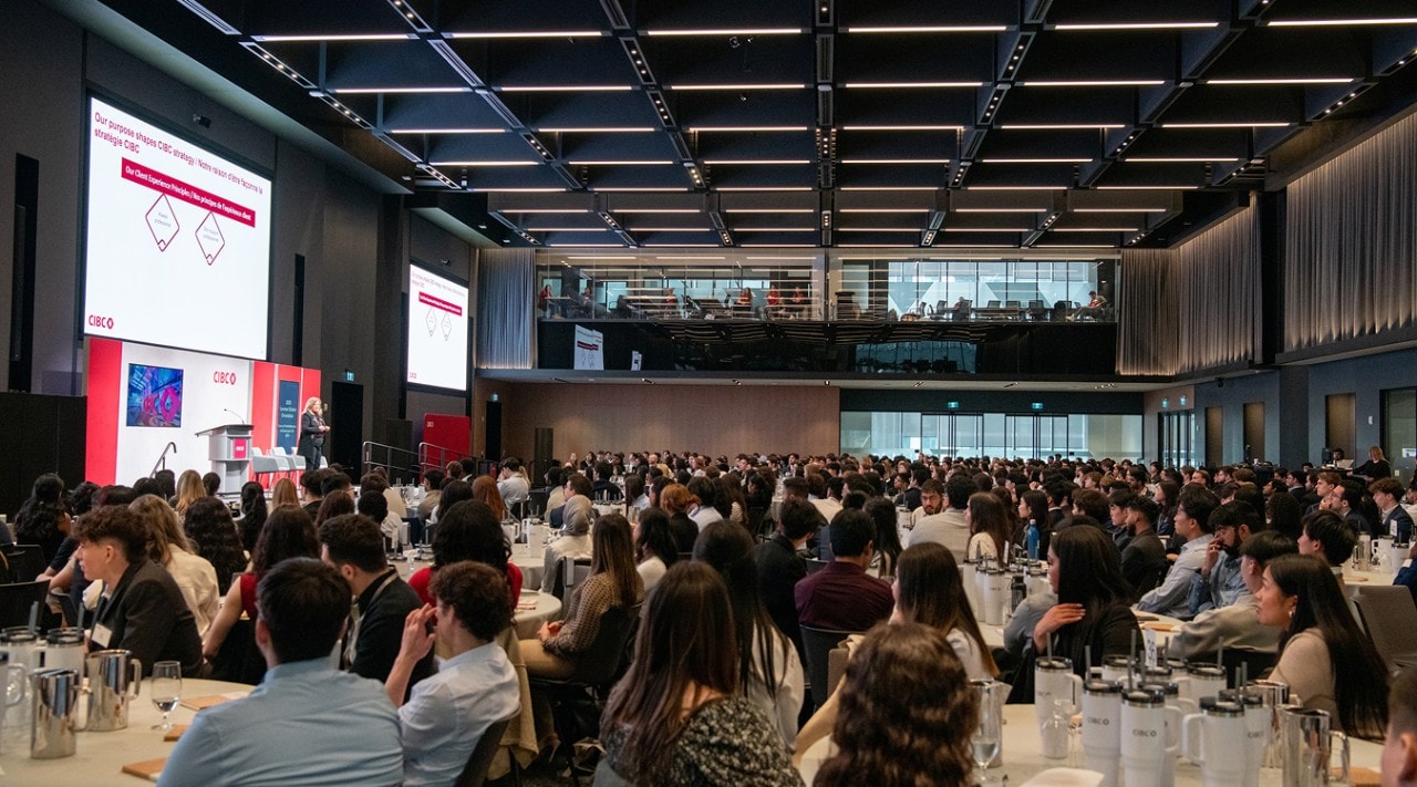 Hundreds of student workshop participants sit at tables in a large conference room and watch a presentation from CIBC.
