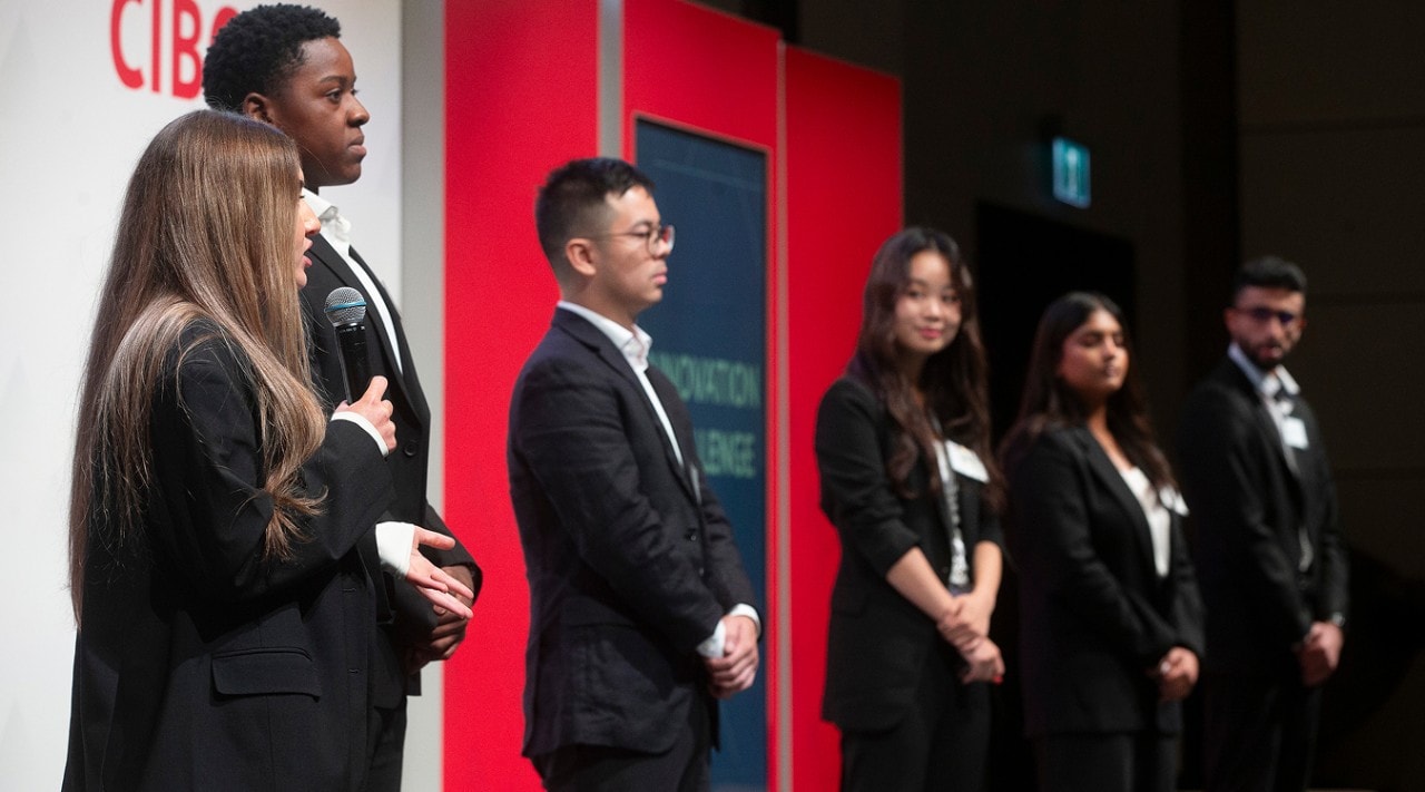 6 students are standing on a stage with the CIBC logo in the background. One student has a microphone and is speaking while the others look on.