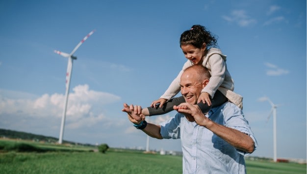 Smiling man with a child on his shoulders walks through a field filled with wind turbines.