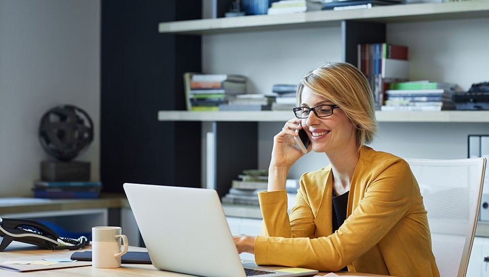 Une femme assise dans son bureau regarde son ordinateur tout en parlant au téléphone.