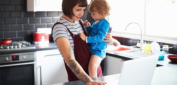 a woman using her laptop while holding her baby in the kitchen