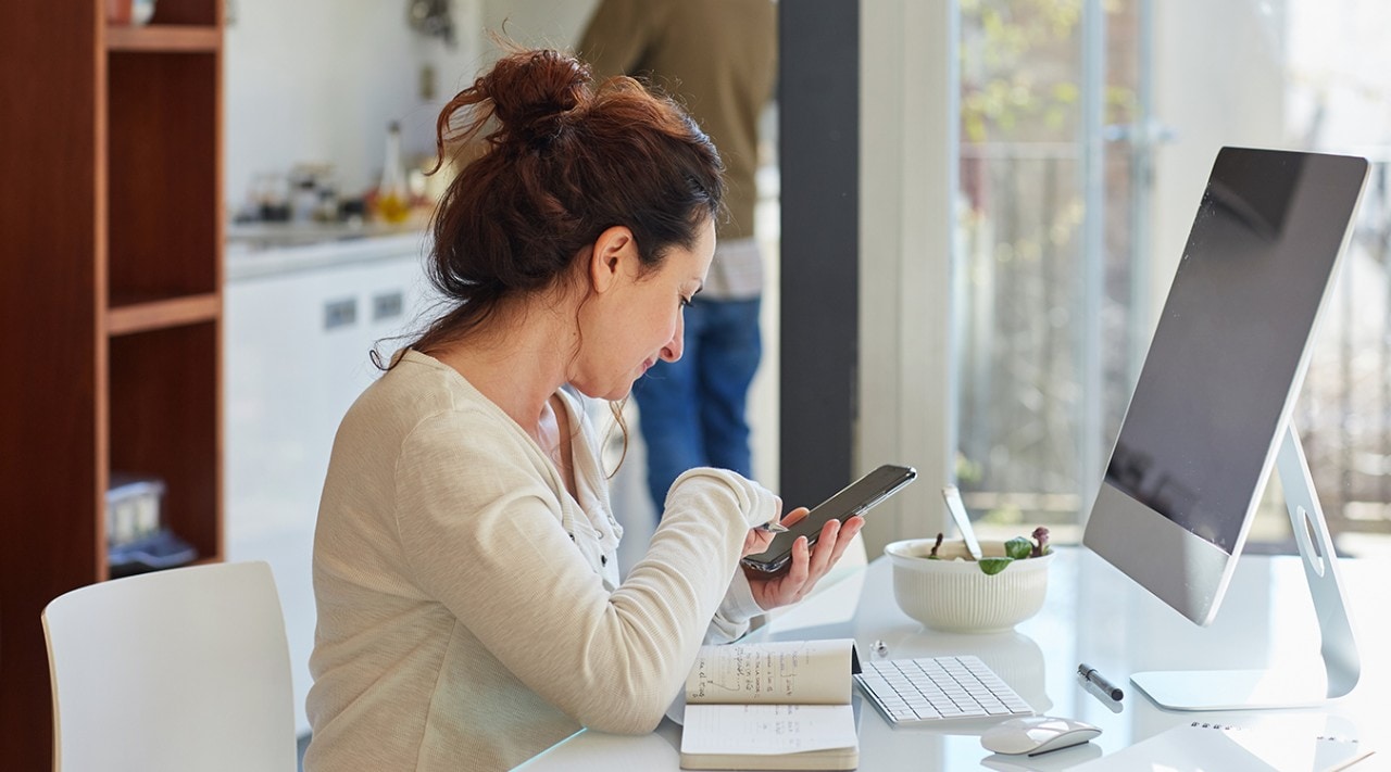 A woman sits at her home computer and checks her phone.