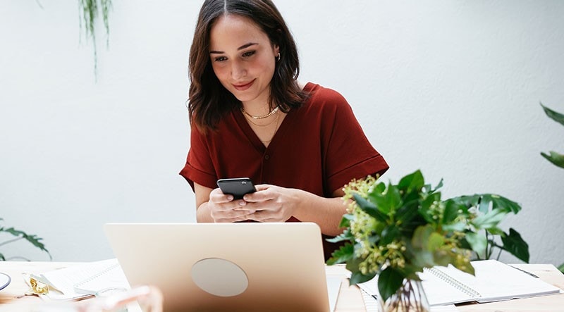 A woman glances at her laptop as she types on her phone.