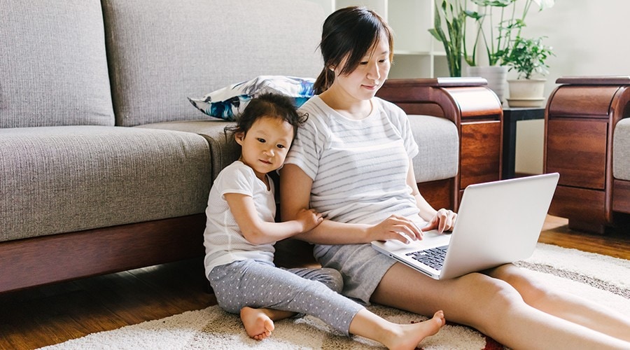 A mother uses her laptop while her child sits beside her