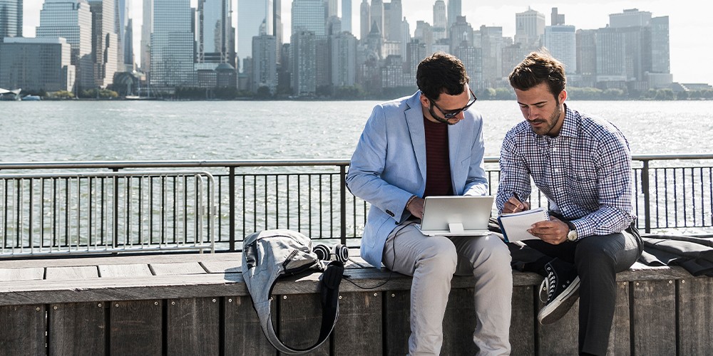 2 hommes d’affaires discutent sur un banc longeant une esplanade avec vue sur Manhattan.