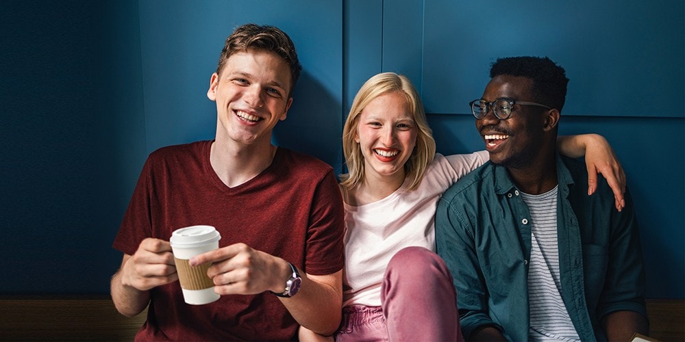 Three young people smiling and enjoying each other’s company. One has a cup of coffee with steam rising from it.